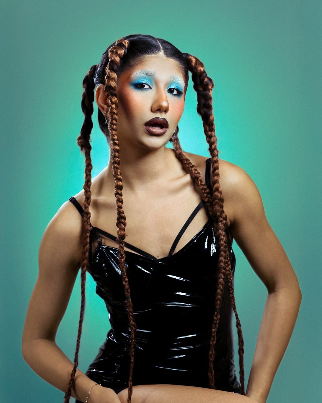 Asal Dabbaghchian in long braids and blue eye makeup, wearing a black vinyl top on a teal-lit music video set