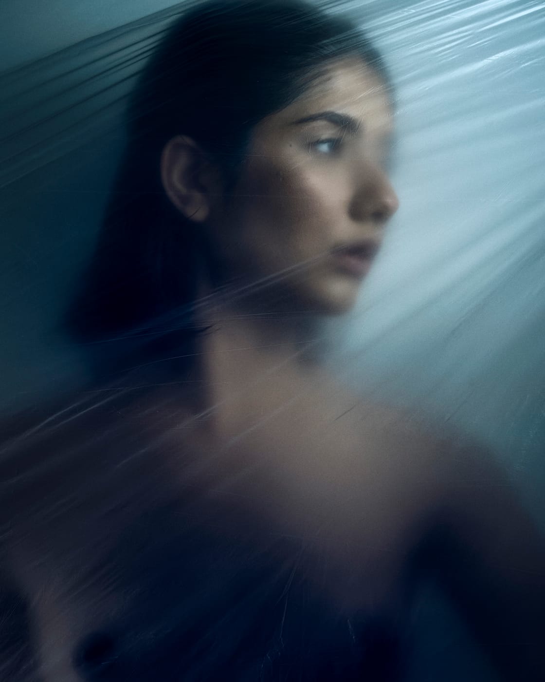Asal Dabbaghchian in profile behind a sheet of clear plastic film, lit in cool blue studio light